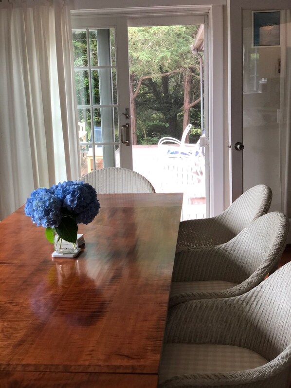 Ein Holztisch mit einer Vase voller blauer Blumen, Korbstühle und Blick durch eine Glastür auf einen Außenbereich mit Bäumen und einer Terrasse.