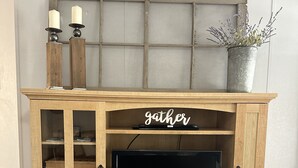 A wooden entertainment center with a flat-screen TV, two candles, and a sign that says "gather". - Peaceful Hill Country Retreat • Private Hot Tub • 3½ Acres • Near Downtown (Wimberley)