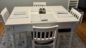 A dining area with a white table set for four, surrounded by white chairs. Above the table, there are three wall-mounted light fixtures. - Peaceful Hill Country Retreat • Private Hot Tub • 3½ Acres • Near Downtown (Wimberley)