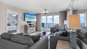 A modern living room with a large flat-screen TV, grey sofas, a coffee table, and a view of the ocean through large windows. - Two Balconies, Sunset/Gulf View, Corner Unit (Destin)