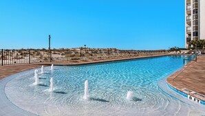 A swimming pool with multiple water jets, a wooden deck, and a sandy beach area. - Two Balconies, Sunset/Gulf View, Corner Unit (Destin)