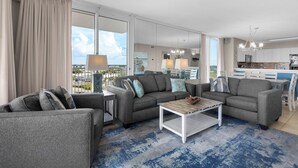 A modern living room with grey sofas, a coffee table, and a view of the cityscape through large windows. - Two Balconies, Sunset/Gulf View, Corner Unit (Destin)