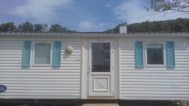 Une maison mobile dotée de deux fenêtres et d’une porte, chacune agrémentée de volets bleus, et d’une petite antenne parabolique fixée au mur.