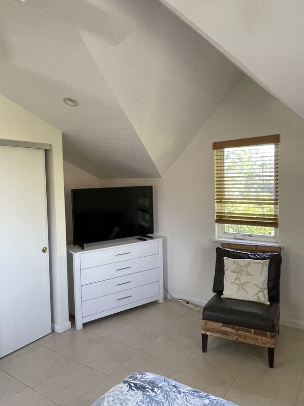 A bedroom with a sloped ceiling, a television on a white dresser, a window with blinds, and a chair with a cushion. - Beautiful Tree Frog Apartment! - South Sound - 5 min walk to Smith Cove (South Sound)