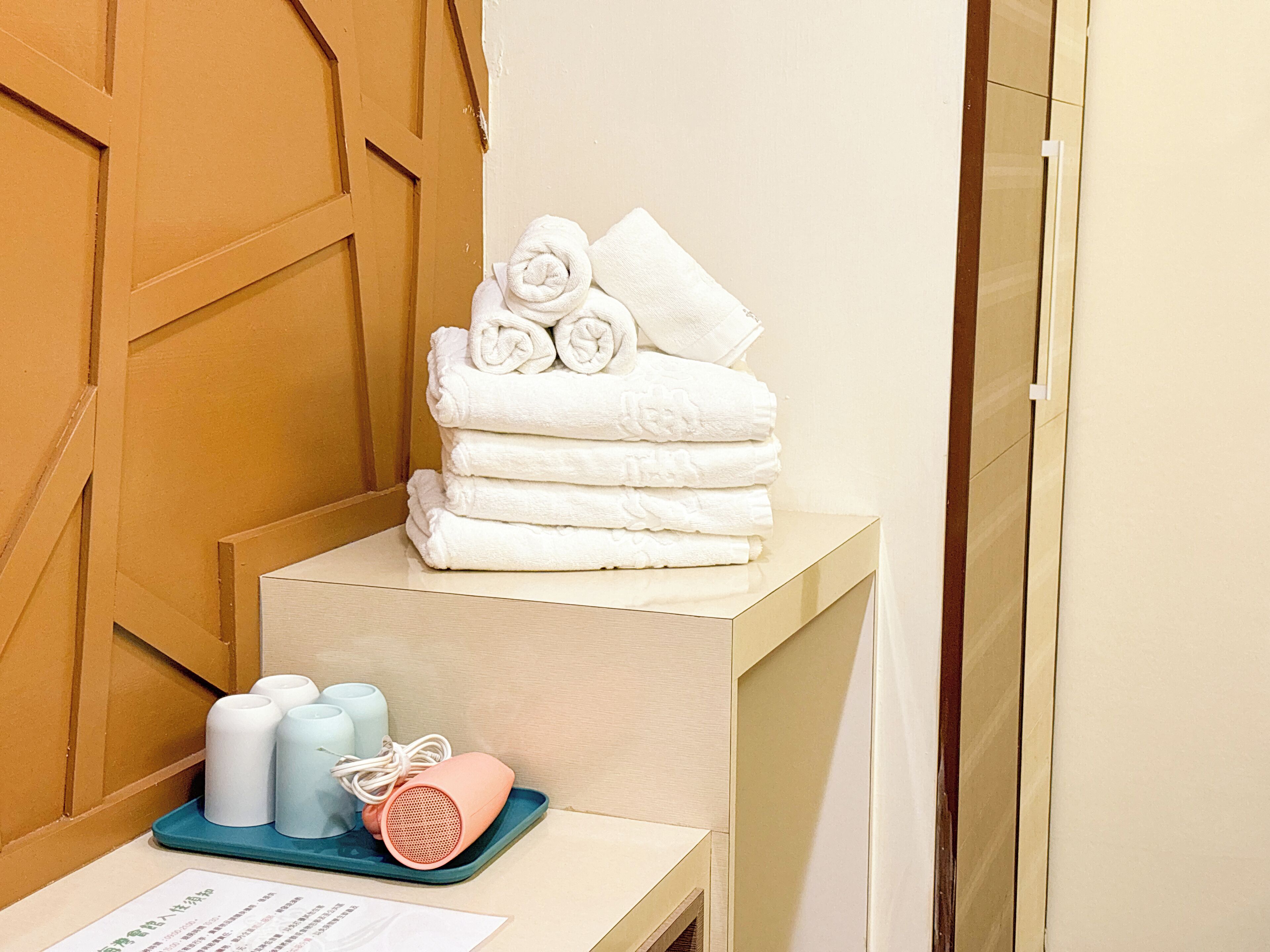 Ein Stapel weiße Handtücher auf einem Podest, eine blaue Ablage mit Toilettenartikeln und ein pinkfarbener Lautsprecher.