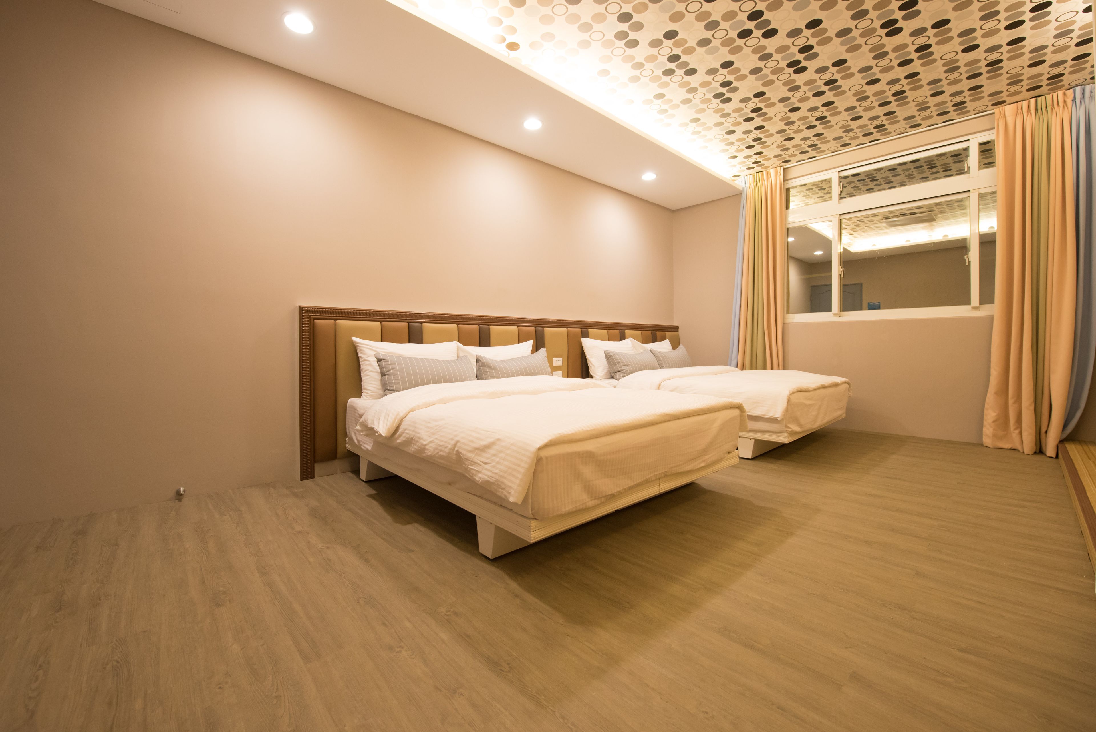 A bedroom with two beds, a wooden floor, a ceiling with circular patterns, and a window with curtains.