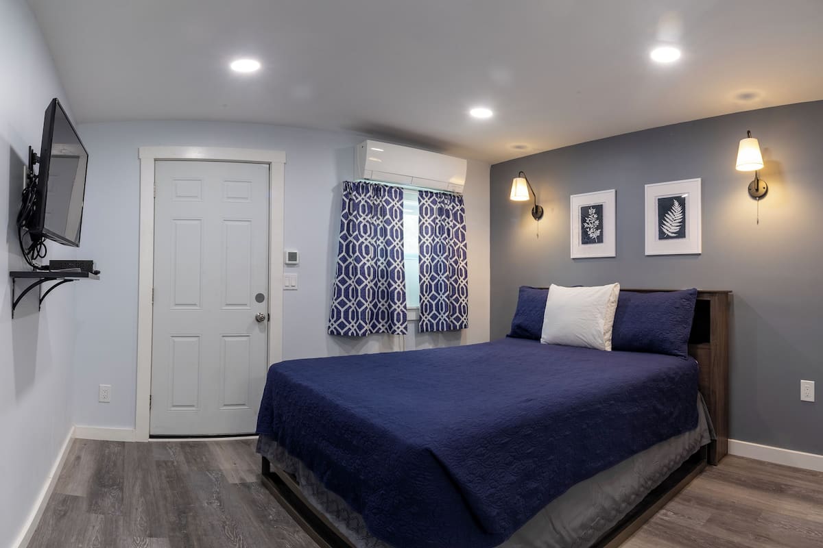A bedroom with a bed, a door, an air conditioning unit, a wall-mounted TV, patterned curtains, and wall-mounted lights.