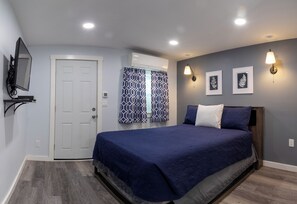 A bedroom with a bed, a door, an air conditioning unit, a wall-mounted TV, patterned curtains, and wall-mounted lights. - Robbins Motel (Bar Harbor)