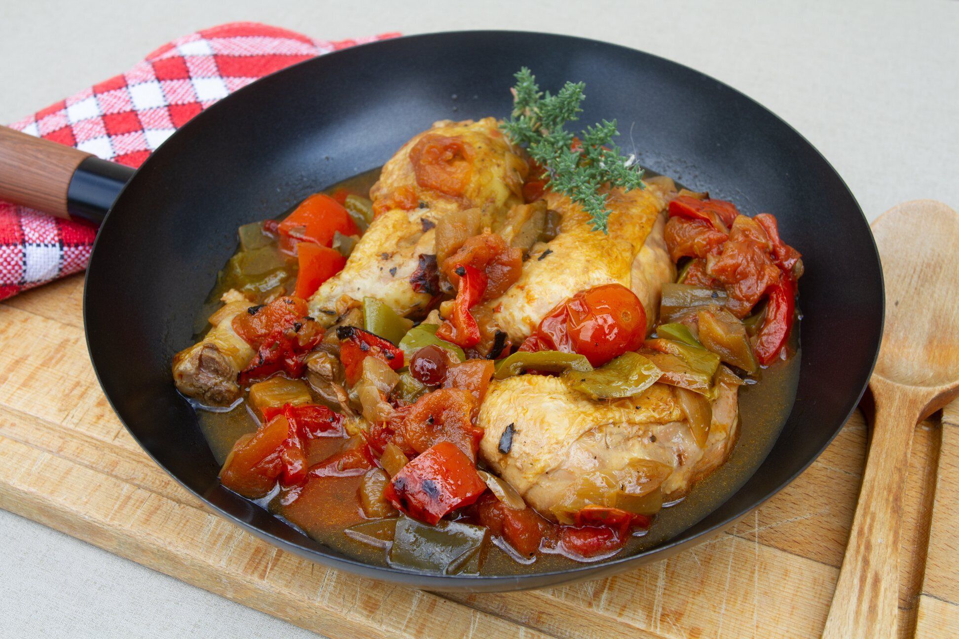 A skillet with chicken and vegetables, possibly a stew or sauté, on a wooden board with a checkered cloth.