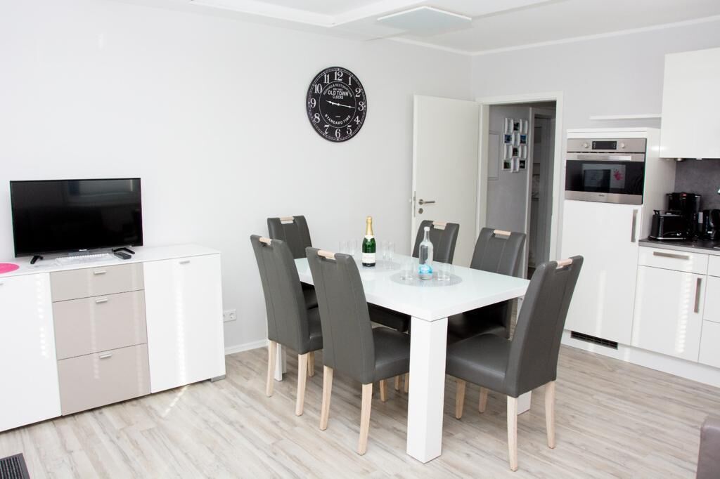 A modern kitchen with white cabinets, a built-in oven, and a dining area with a white table and grey chairs.