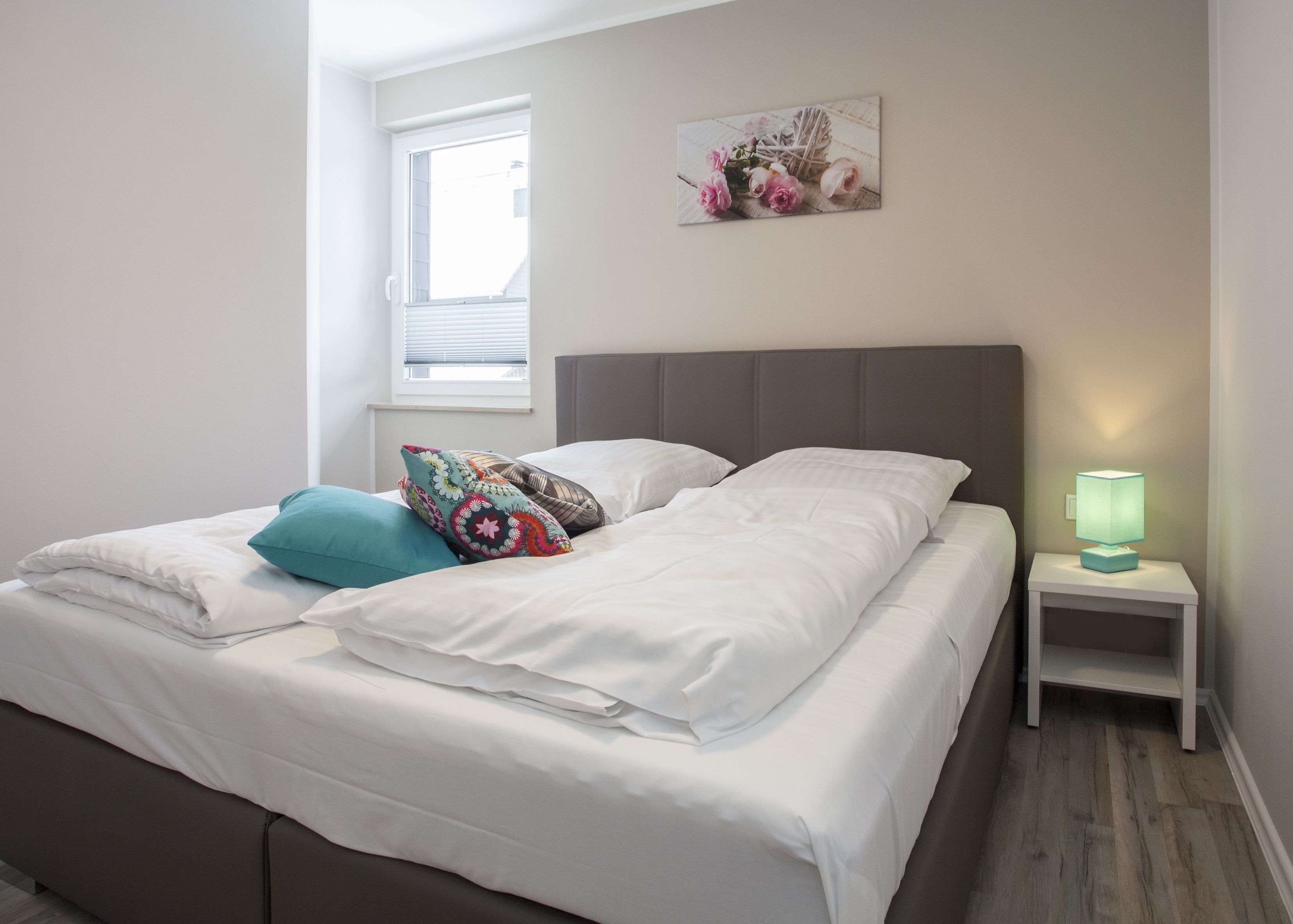 A neatly made double bed with white linens, a teal pillow, and a colorful patterned pillow. There is a small bedside table with a green lamp, and a window with blinds to the left.