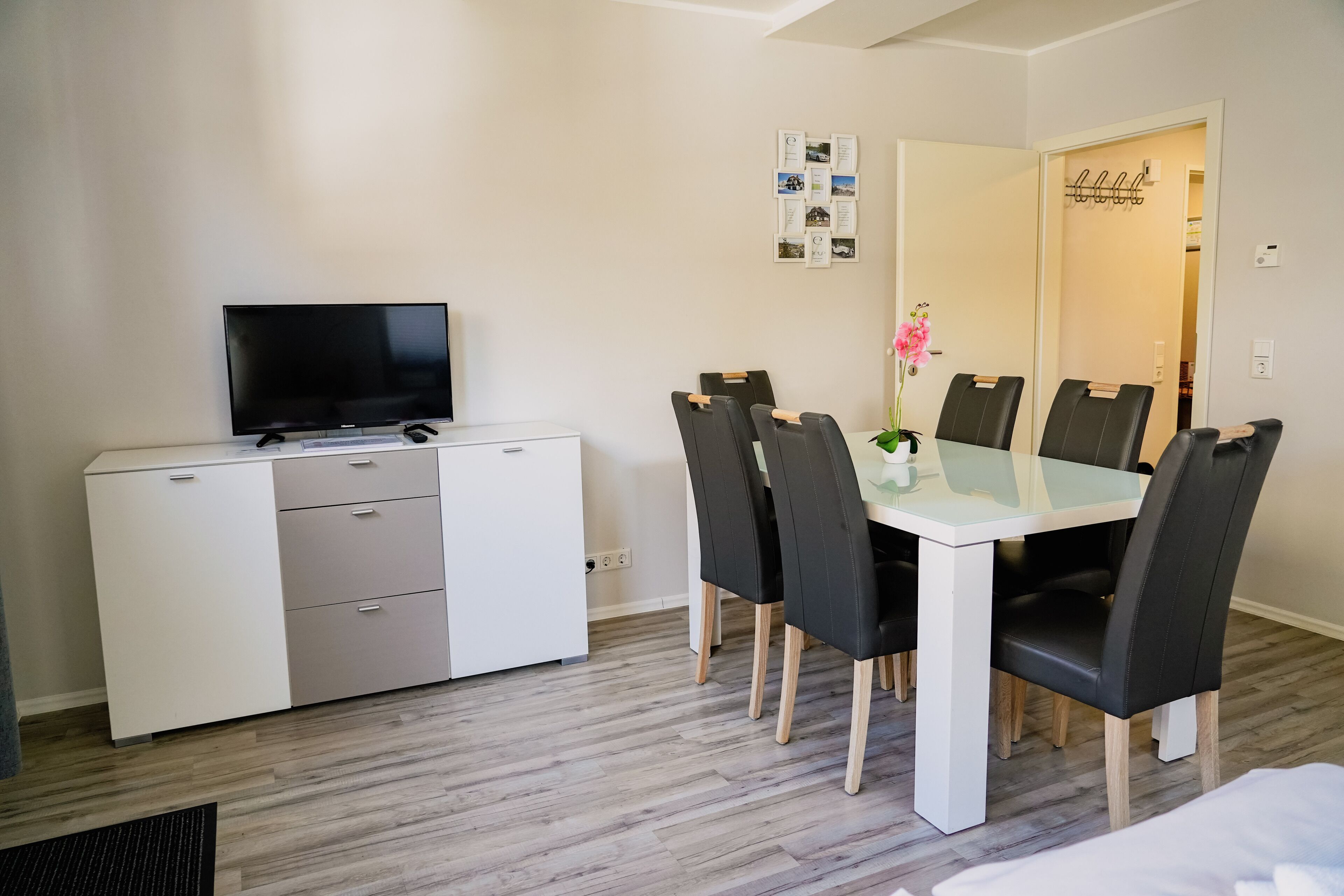 A modern room with a flat-screen TV on a white cabinet, a dining table with black chairs, and a small vase with pink flowers.