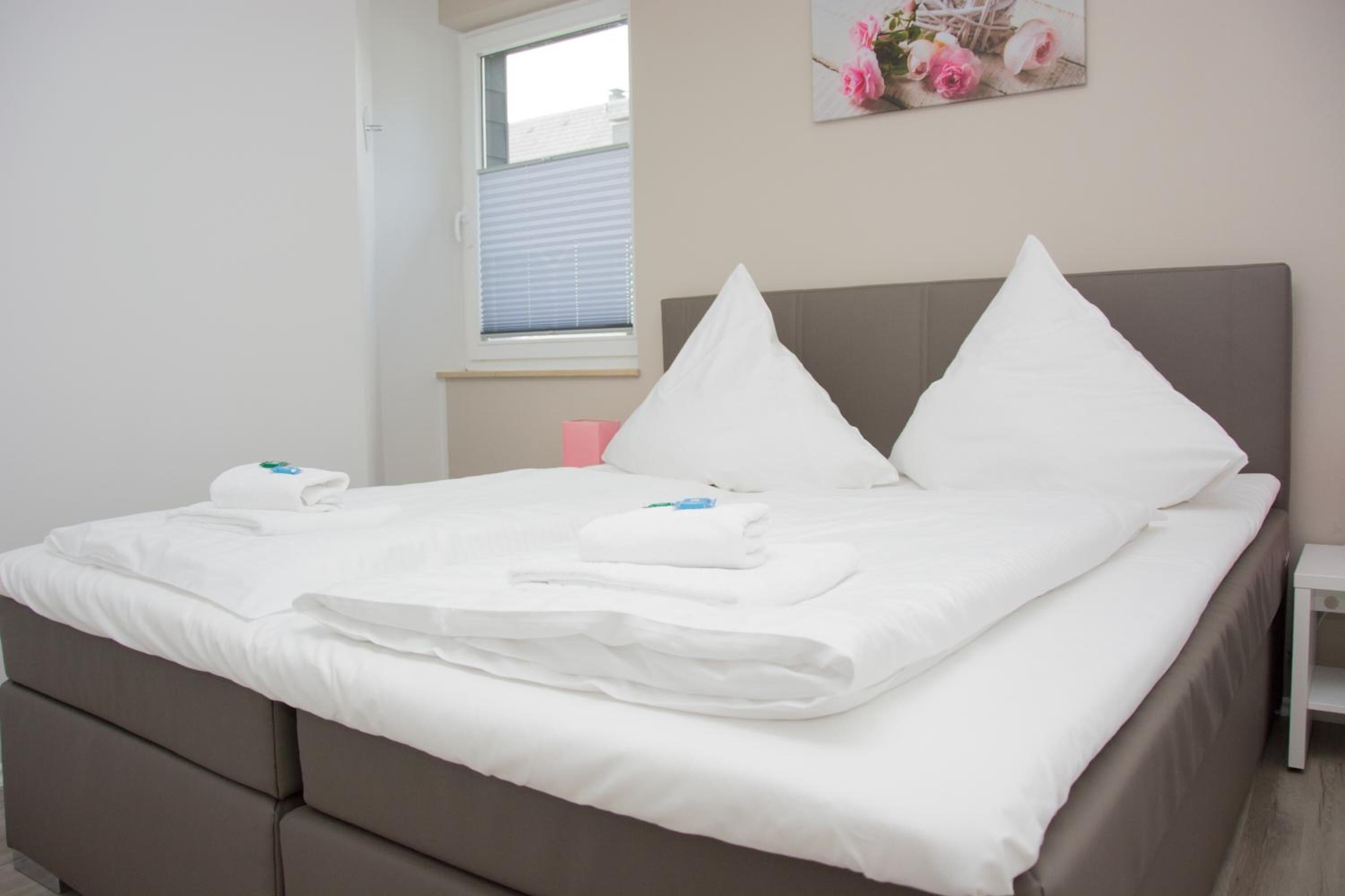 A neatly made bed with white linens and pillows, a grey headboard, and a small bedside table with a pink object on it.