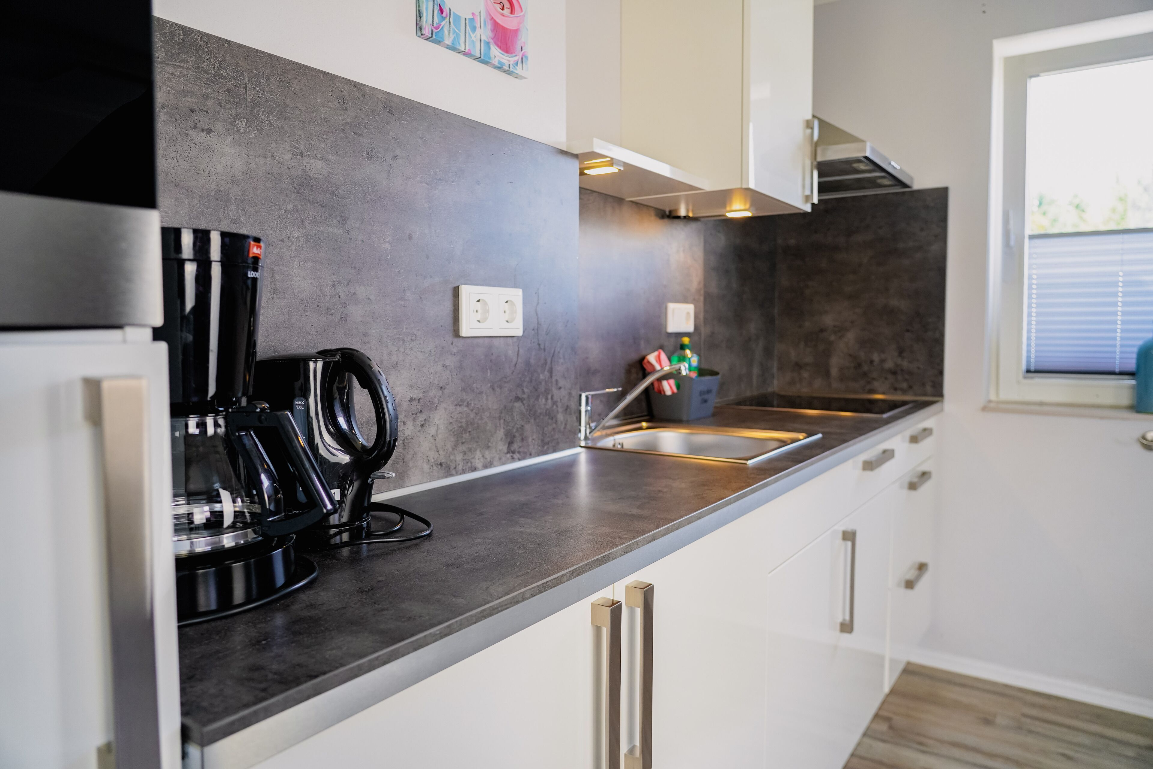 A modern kitchen with a stainless steel countertop, a coffee machine, and a window with blinds.