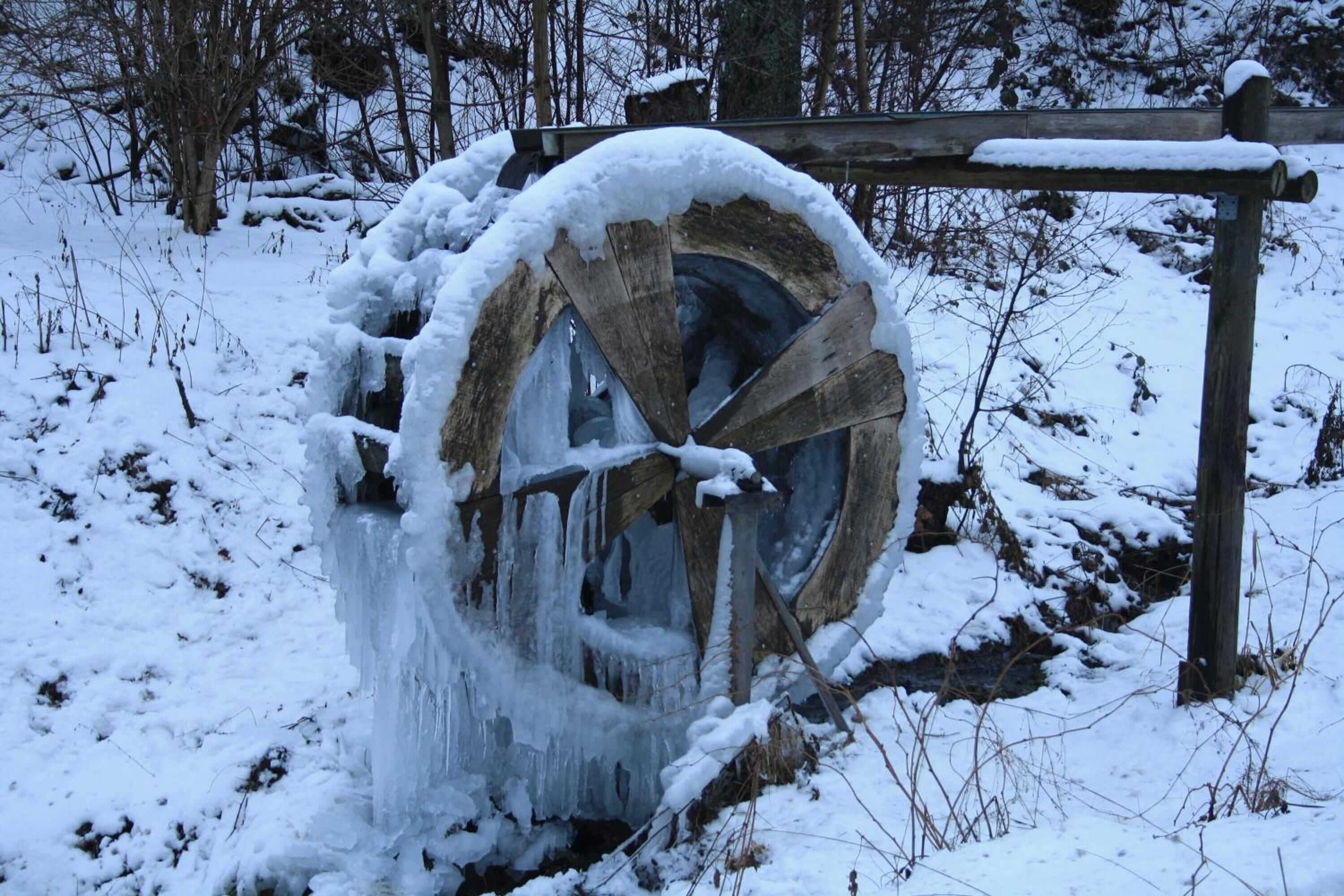 A wooden water wheel partially covered in snow and ice, surrounded by a snowy landscape.