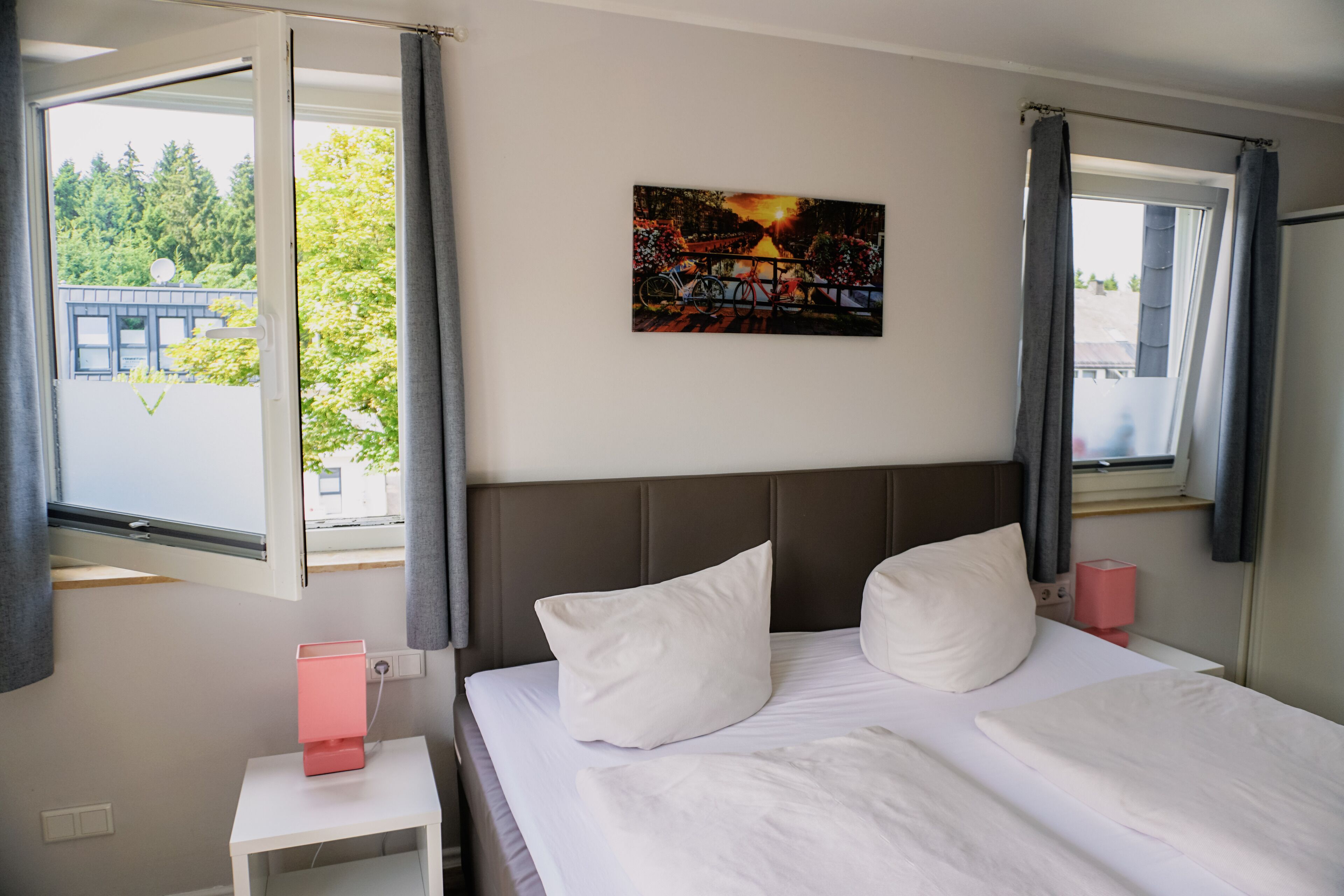 A hotel room with a bed, a nightstand, a painting, and a window with curtains.