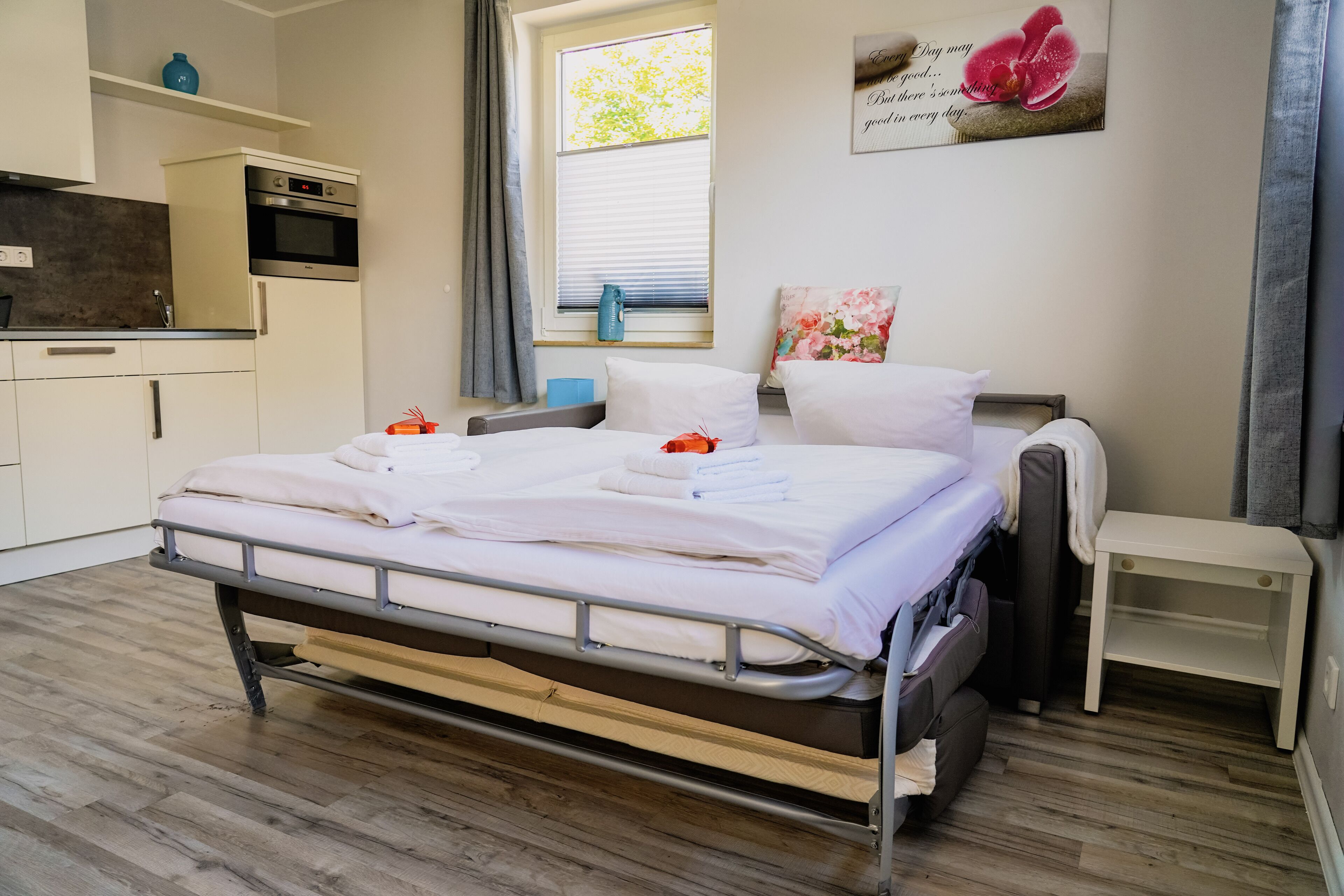 Een net opgemaakt bed met witte lakens en handdoeken, een klein nachtkastje, een raam met zonwering, en een keukenhoek met een fornuis en kastjes.