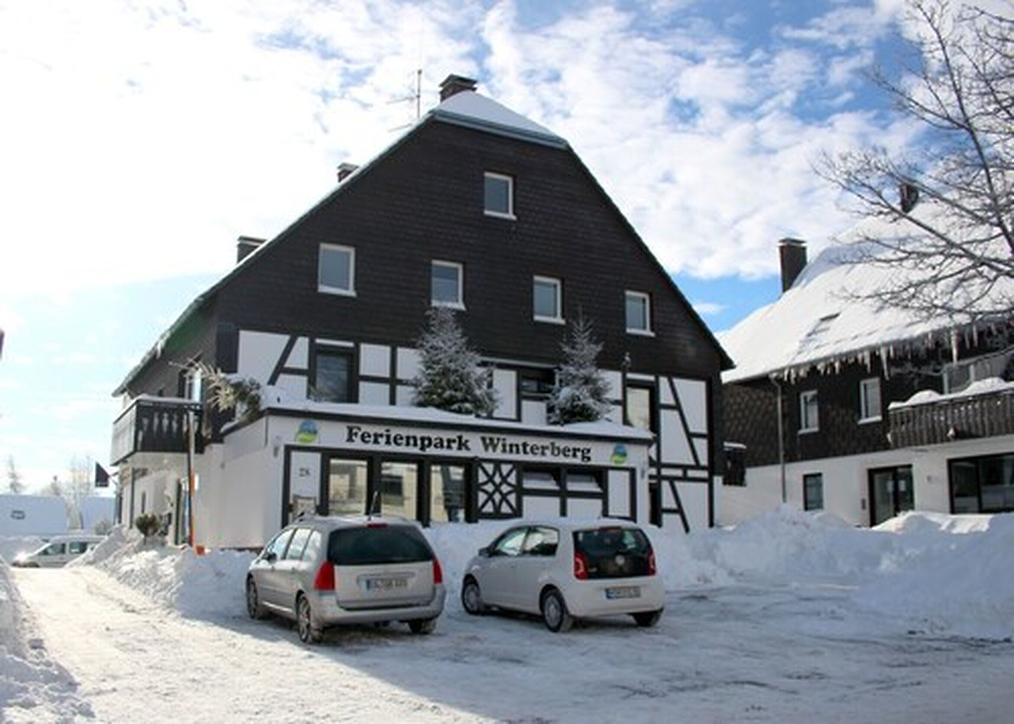 Un edificio llamado 'Ferienpark Winterberg' con fachada de entramado de madera tradicional, coches estacionados y nieve en el suelo.