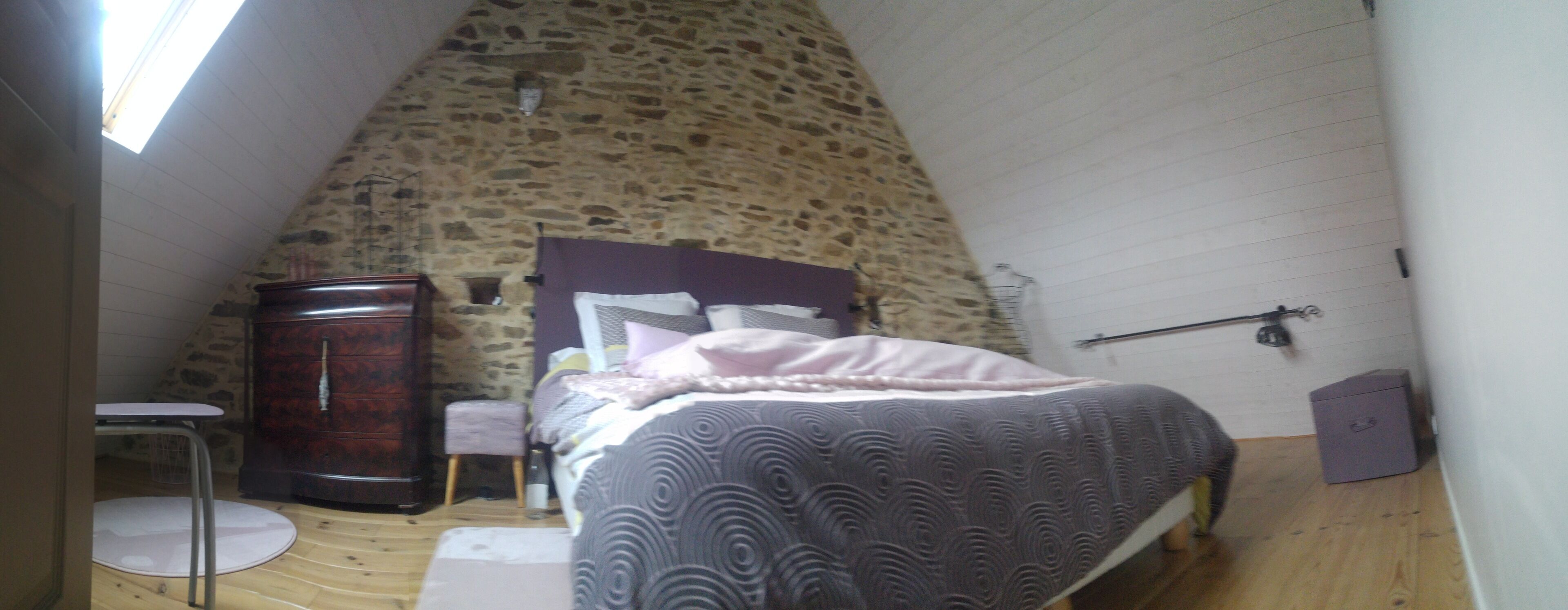 A bedroom with a sloped stone ceiling, a bed with a patterned bedspread, a wooden dresser, and a small round table.