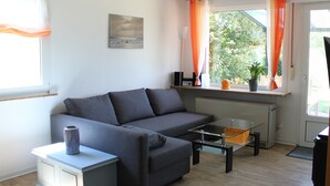 Un salon moderne avec un canapé gris, une table basse en verre et un petit meuble. On y trouve une fenêtre avec des rideaux orange, un radiateur et une plante en pot sur le rebord de la fenêtre.