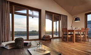 Une pièce spacieuse dotée de grandes portes-fenêtres coulissantes donnant sur un espace extérieur, une table à manger en bois préparée pour deux personnes et un coin lecture confortable avec des livres et une lampe.
