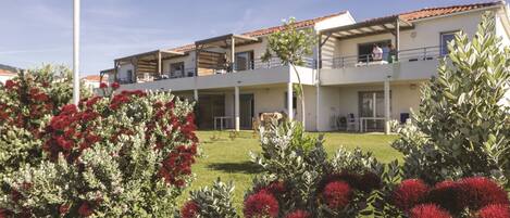 A multi-story building with balconies, a flat roof, and a well-maintained garden with red flowers.