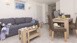A modern living room with a gray sofa, a wooden coffee table, and a dining area with a table set for two. - Résidence Odalys Acqua Bella (Poggio-Mezzana)