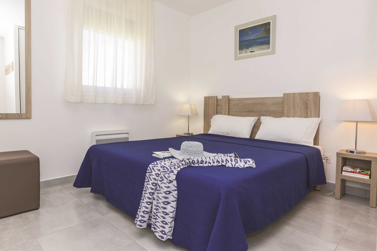Un dormitorio ordenado con una cama grande, dos mesitas de noche con lámparas, una mesita de noche pequeña con un libro, un cuadro en la pared y una ventana con cortinas translúcidas.