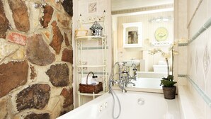 A bathroom with a stone wall, a white bathtub, a white sink, and a mirror. There are shelves with baskets and toiletries. - Romantic, 1 bedroom cottage walking distance to downtown, pet friendly, private. (Eureka Springs)