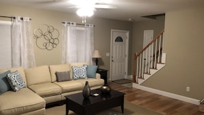 A living room with a beige sofa, a coffee table, and a brown leather chair. There is a staircase with wooden railings leading to a door. - Immaculate Cape Cod Family Retreat- minutes from Ponds, Lakes, and Ocean! (Sandwich)
