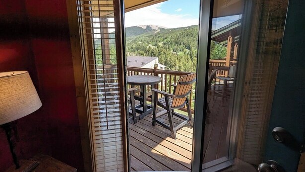Une terrasse en bois avec une table et des chaises, une vue sur un paysage montagneux et une porte dotée d’un panneau en verre.