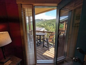 Una terraza de madera con una mesa y sillas, y vistas a un paisaje montañoso, además de una puerta con un panel de vidrio.
