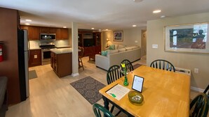 A modern kitchen with stainless steel appliances, a dining area with a wooden table and chairs, and a living area with a sofa and TV.