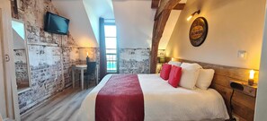 Ein Schlafzimmer mit Backsteinwand, einem Bett mit roten und weißen Kissen, einem Schreibtisch mit Stuhl und einem an der Wand befestigten Fernseher.