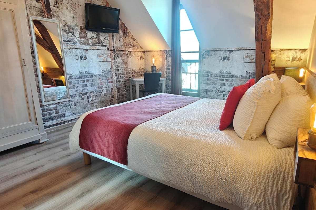 Ein Schlafzimmer mit schrägverdeckter Decke, freigelegtem Mauerwerk, einem großen Bett mit roter Decke, einem hölzernen Nachttisch mit Kerze und einem an der Wand montierten Fernseher.
