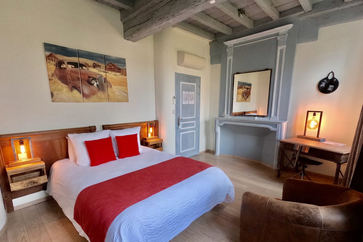 Ein Schlafzimmer mit Bett, Holz-Kopfteil, Bettwäsche in Rot und Weiß, einem braunen Ledersessel, einem Schreibtisch mit Lampe und einem Bild an der Wand.