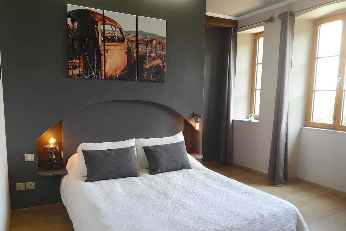 Ein modernes Schlafzimmer mit einem großen Bett, einem markanten, gebogenen Kopfteil, einem Wandbild und einem Fenster mit Vorhängen.