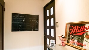 Un área de recepción con una pizarra mostrando un mensaje de bienvenida y un reloj. Hay una puerta con un ventanal, y un mostrador con un cartel enmarcado y dos figuritas.