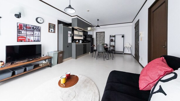 Una sala de estar moderna con un televisor de pantalla plana, un sofá negro y una mesa de centro redonda. En el fondo, se observa una zona de cocina con una mesa de comedor y sillas.