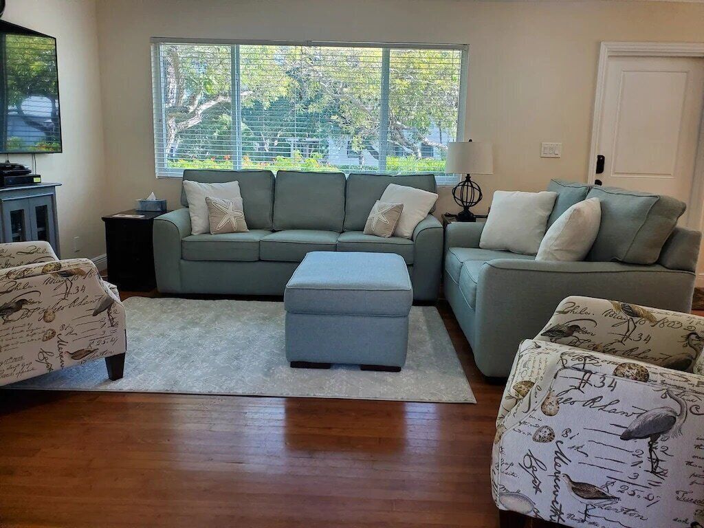 A living room with a sectional sofa, ottoman, and patterned armchairs, a flat-screen TV, and a window with a view of trees.