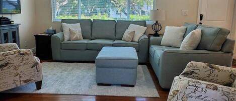 A living room with a sectional sofa, ottoman, and patterned armchairs, a flat-screen TV, and a window with a view of trees.