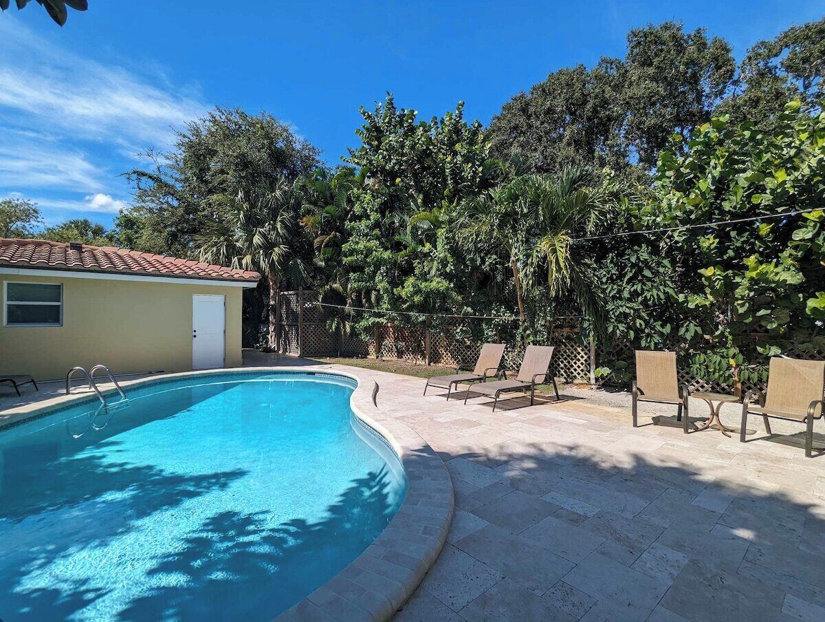 A swimming pool with a tiled deck, surrounded by trees and a building with a red roof.