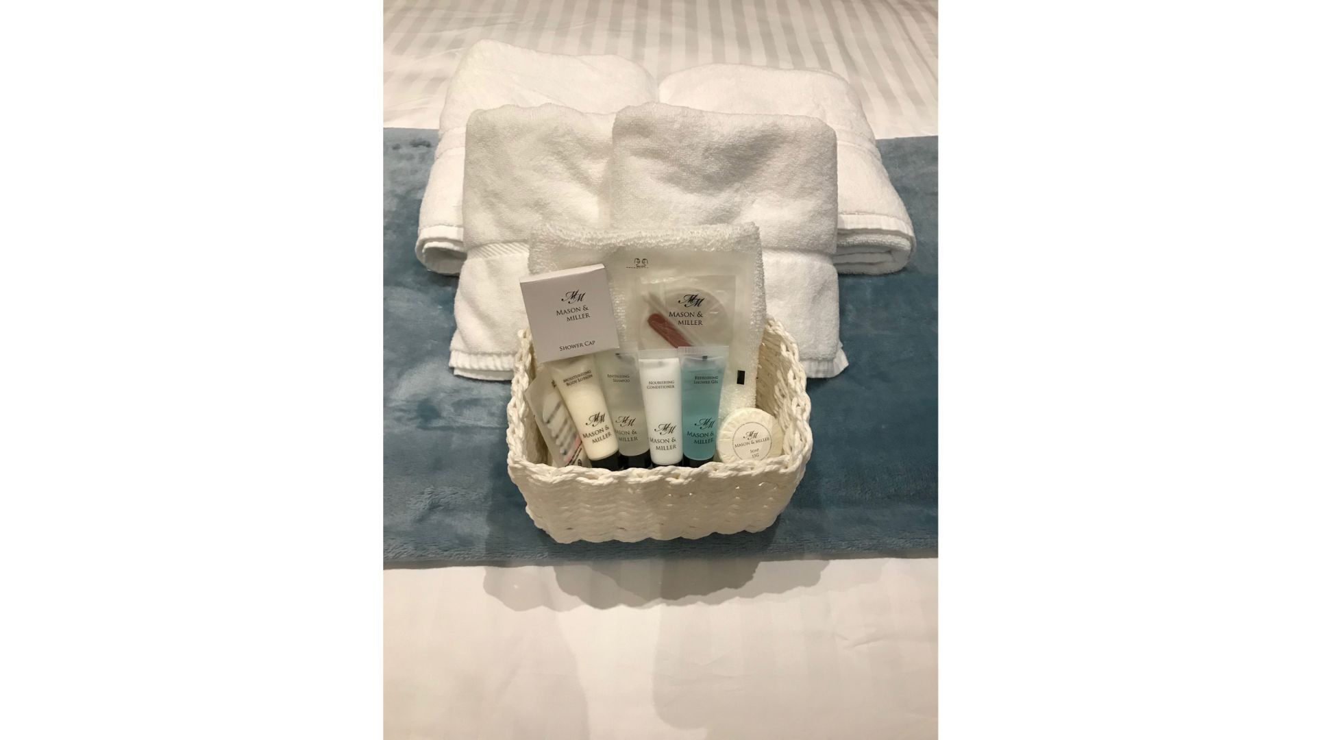 A basket of toiletries including a shower cap, moisturizing lotion, and a soap bar, placed on a bed with folded towels.