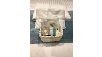 A basket of toiletries including a shower cap, moisturizing lotion, and a soap bar, placed on a bed with folded towels.