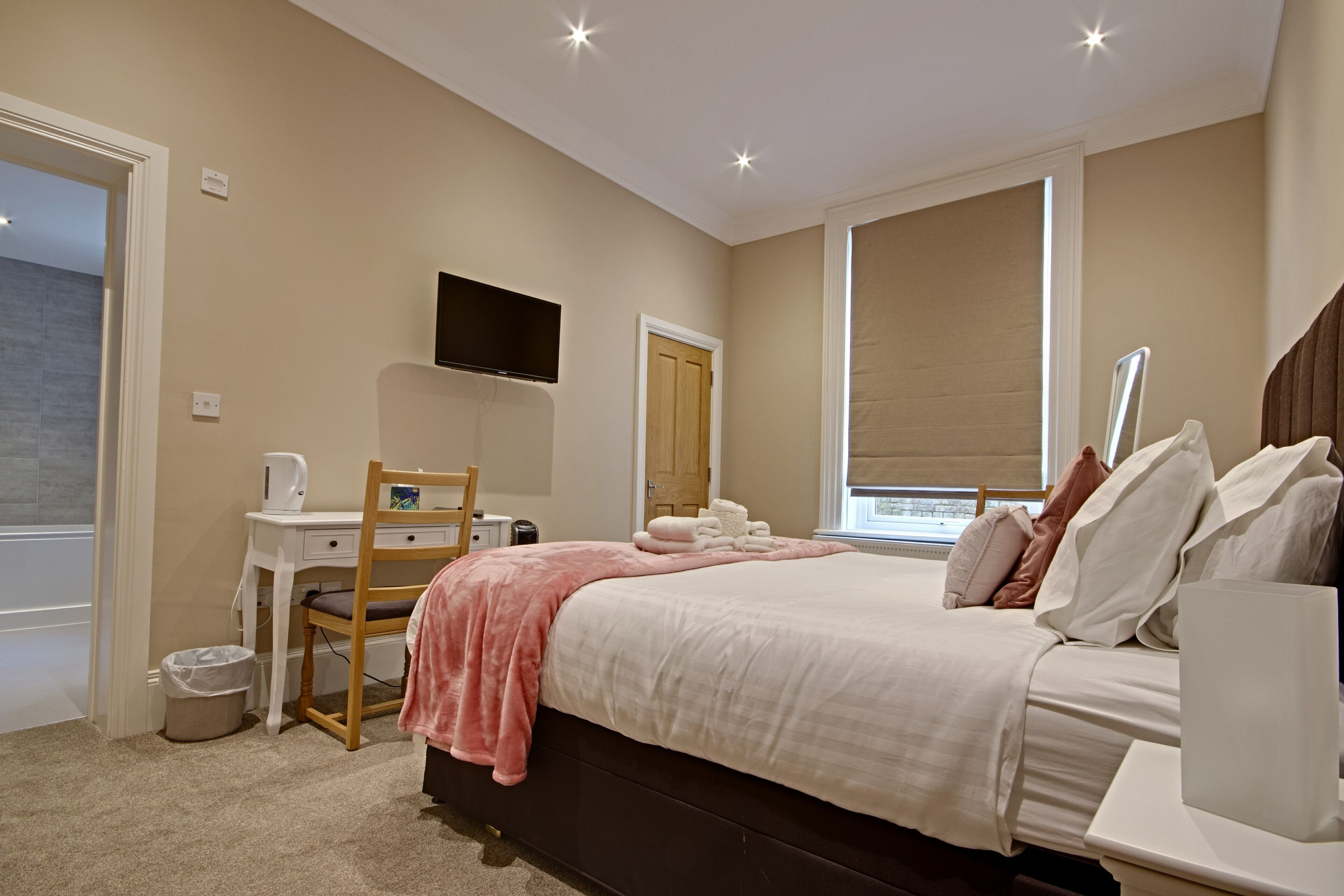 A bedroom with a bed, a desk, a chair, a television, and a window with blinds.