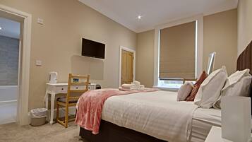 A bedroom with a bed, a desk, a chair, a television, and a window with blinds.
