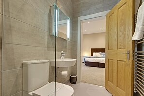 Un baño moderno con inodoro, lavamanos y espejo. Una puerta de madera da acceso a un dormitorio con cama y mesita de noche.