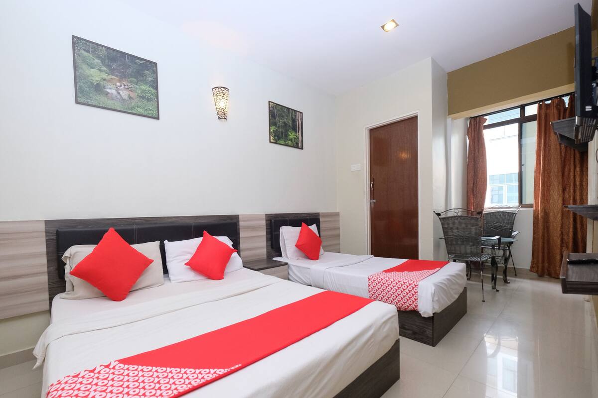 A hotel room with two beds, each with red pillows and a red and white patterned bedspread. There are two wall-mounted light fixtures and two framed pictures on the wall.