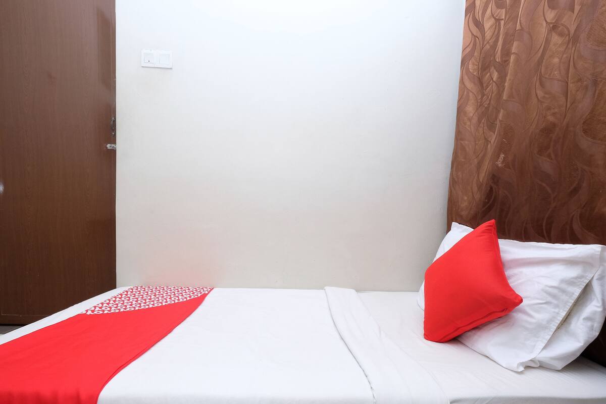 A single bed with a white and red bedspread, a red pillow, and a brown curtain.