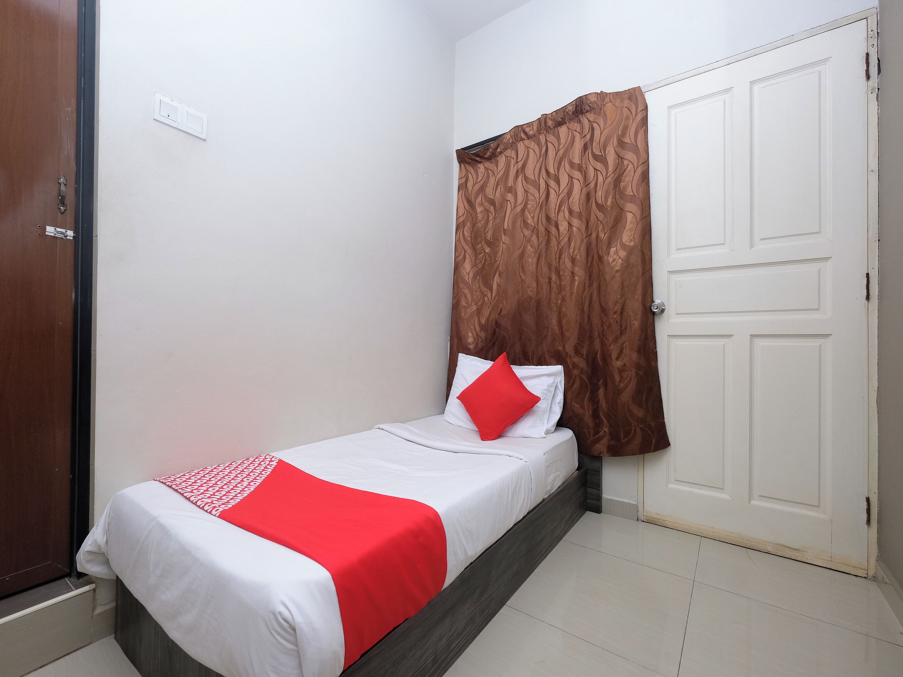A small, neatly made bed with white and red bedding, a brown curtain, and a white door in a simple room.